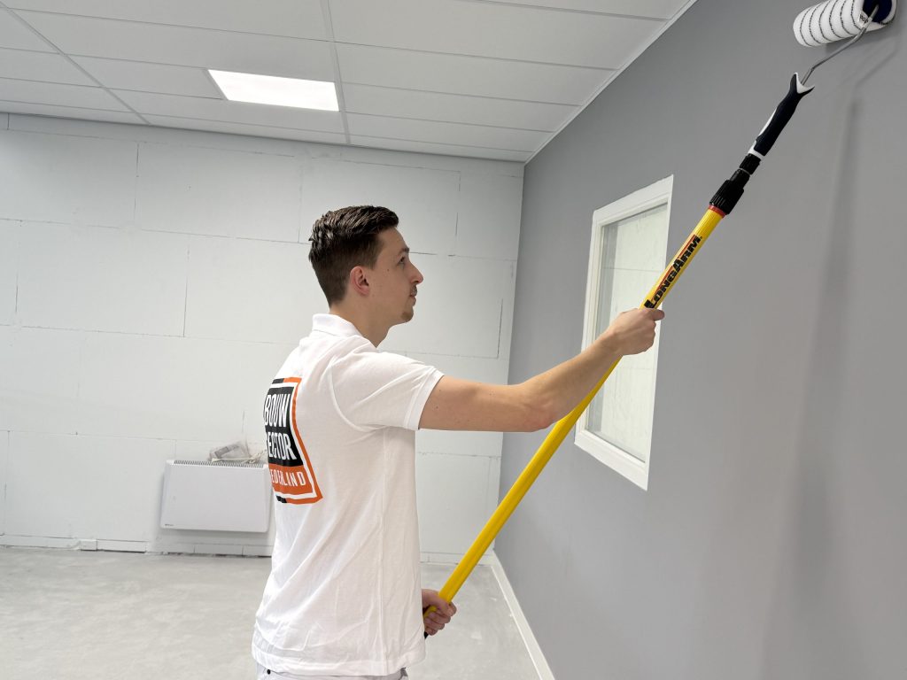 Schilder rolt een grijze wand naast een raam in een wit gelakt kozijn
