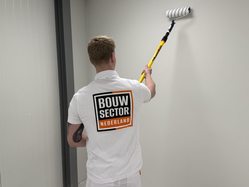 Schilder met het logo van Bouwsector Nederland op zijn rug rolt een lichte muur tot aan het plafond