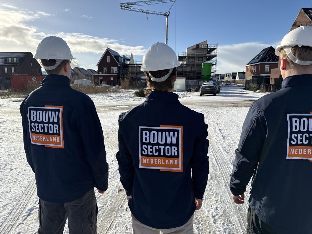 Drie vakmensen van Bouwsector Nederland in winterjassen kijken naar rijtjeshuizen met bakstenen gevels
