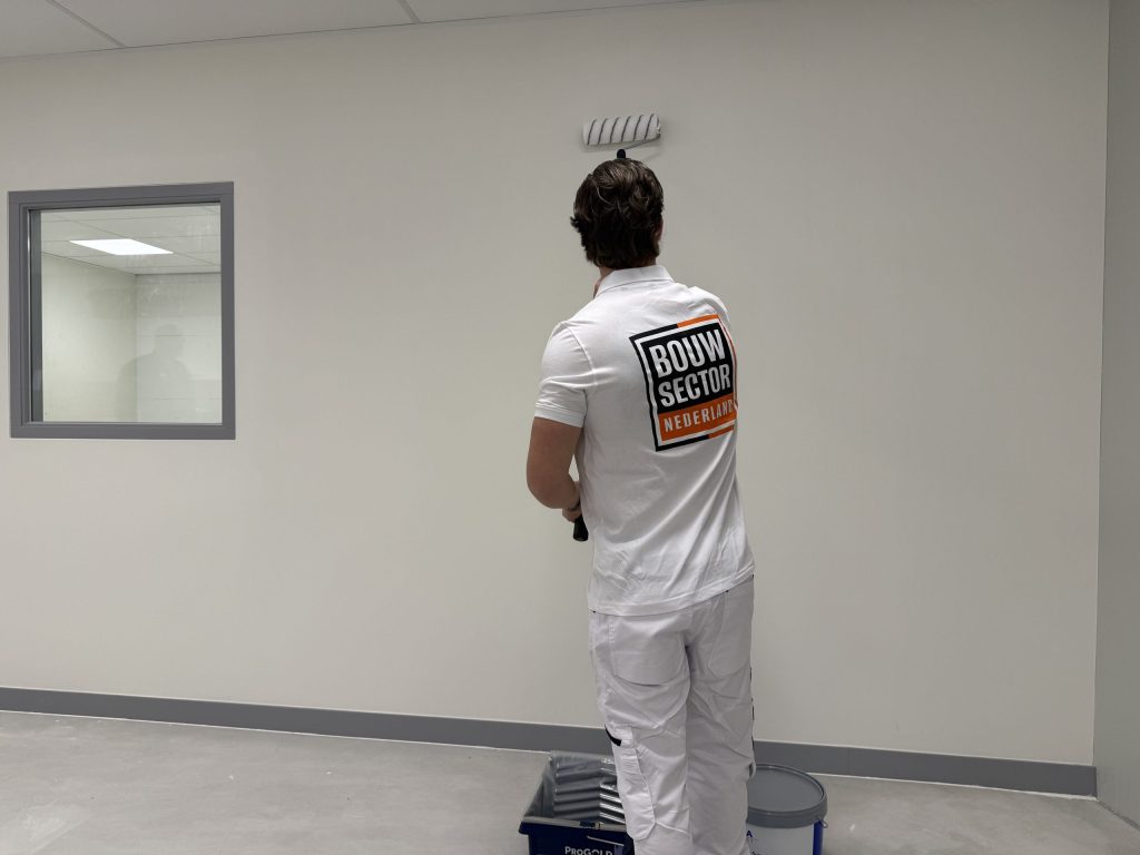 Schilder in wit werktenue rolt een lichte wand tot onder het systeemplafond, met verfbak en emmer op de vloer