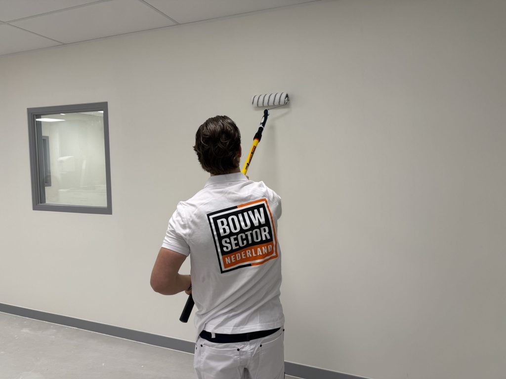 Schilder van Bouwsector Nederland rolt een witte wand met een roller aan een gele steel, naast een binnenraam