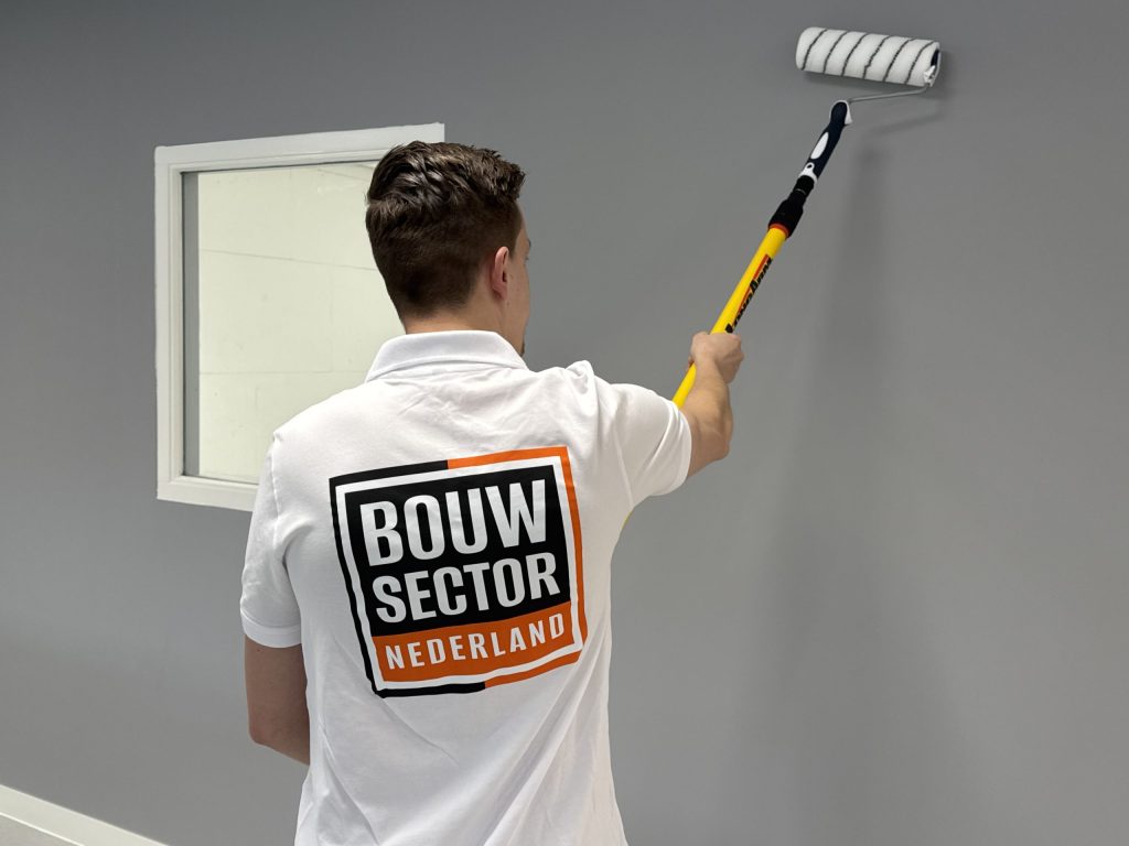 Schilder met het Bouwsector Nederland-logo rolt een grijze wand naast een raam met wit kozijn