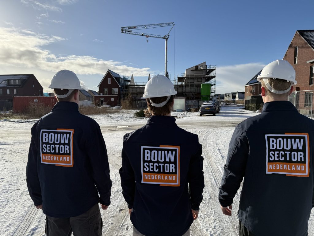 Drie vakmensen van Bouwsector Nederland in werkjas met witte helm, gezien op de rug, bij een woonwijk in aanbouw in de sneeuw